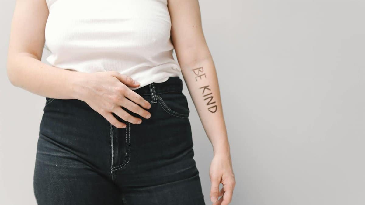 Person in jeans and white tank top with 'Be Kind' text on arm, promoting kindness.