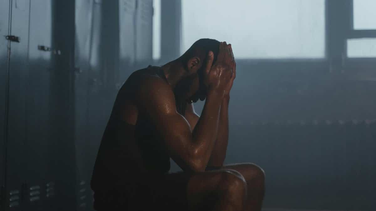 Sweaty man resting on bench in dimly lit locker room, showing exhaustion and recovery.