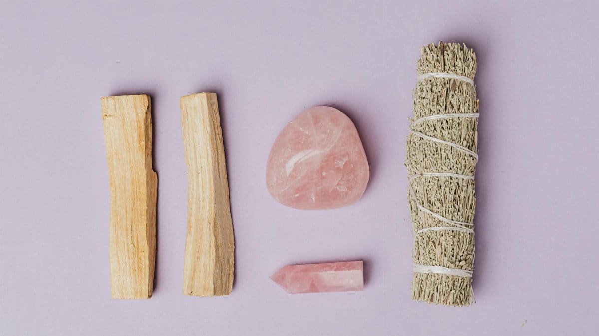 Flat lay of rose quartz, sage, and palo santo on a purple surface, ideal for healing themes.