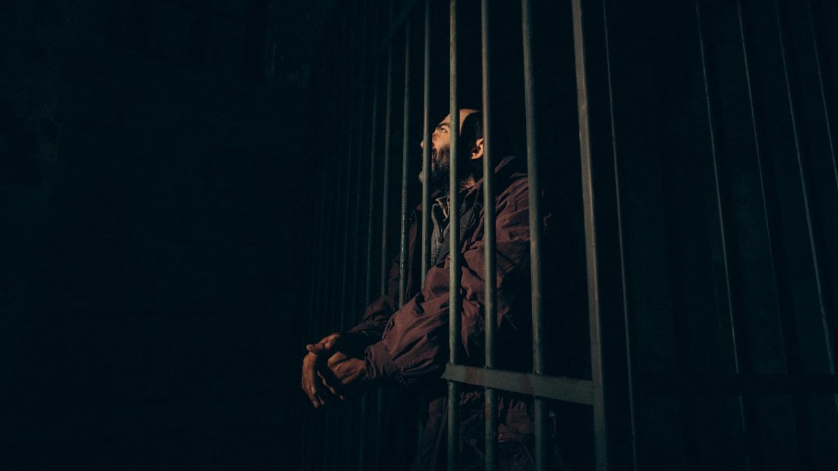 A man stands behind bars in a dark prison cell, evoking a sense of confinement and solitude.