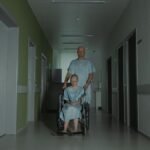 Two patients in hospital gowns navigate a hallway, symbolizing struggle and resilience.