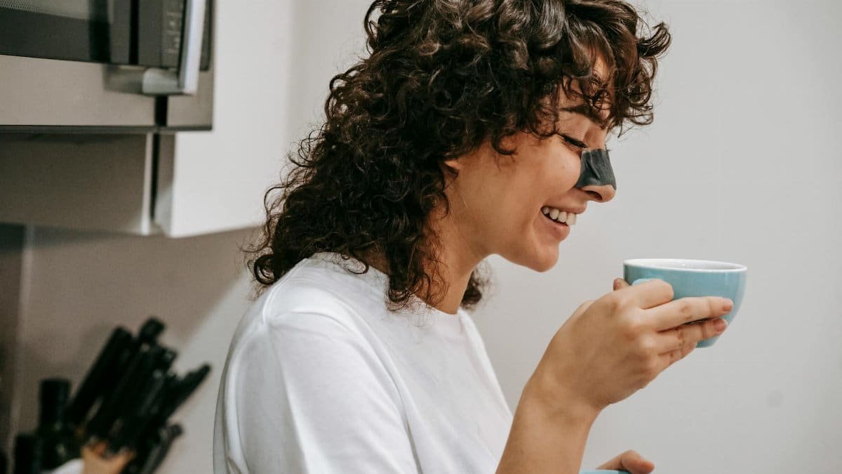 Woman with curly hair enjoys a hot beverage in the kitchen, wearing a nose strip.