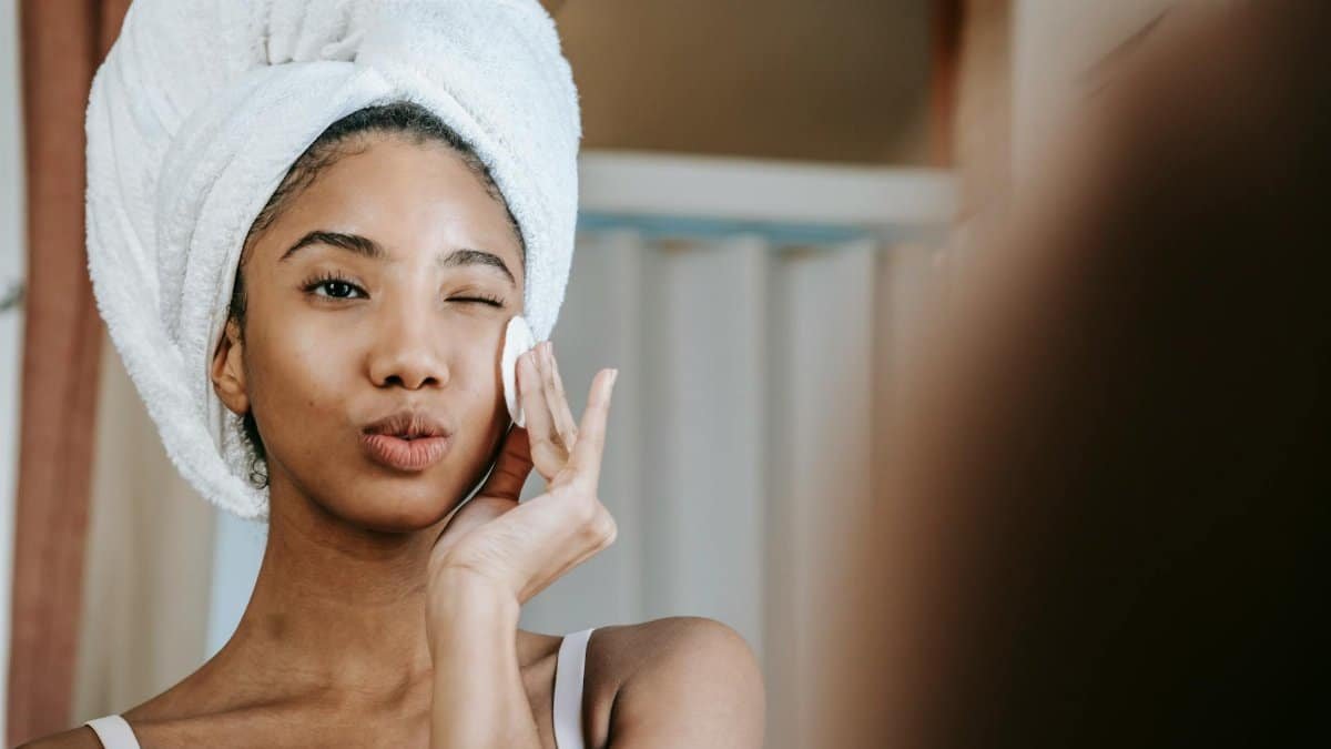Charming ethnic female with folded lips winking while cleaning face with cotton pad during daily beauty routine