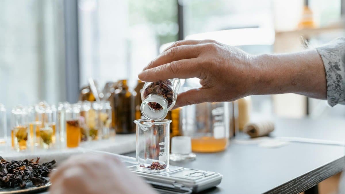 Detailed view of hands adding dried botanicals to a glass beaker in a lab setting.