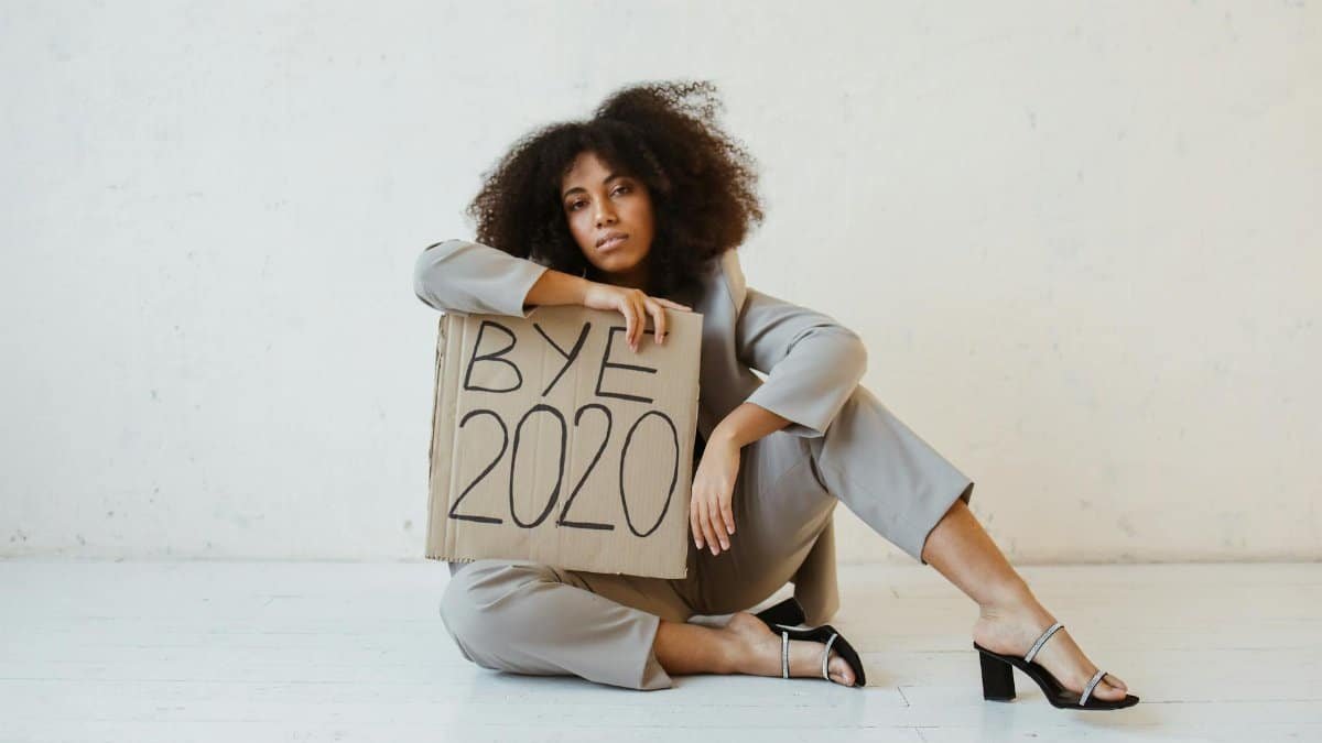 Elegant woman in gray suit sitting indoors holding a 'Bye 2020' sign, symbolizing reflection and hope.