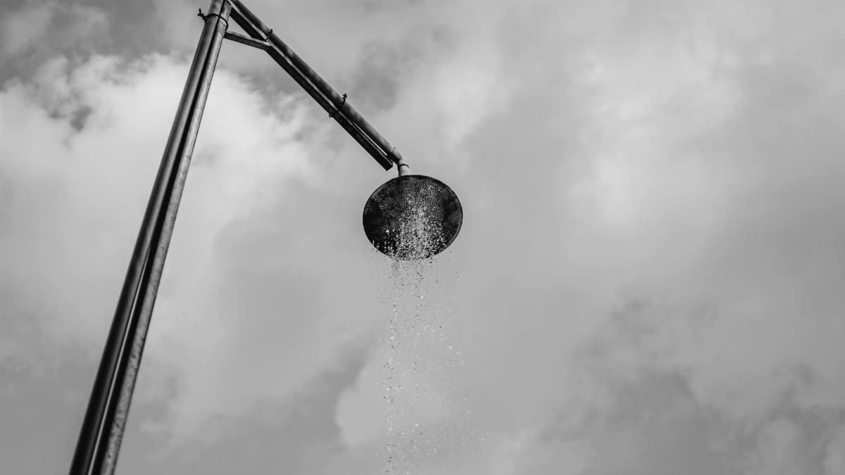 A black and white depiction of an outdoor shower with water against a cloud-filled sky.