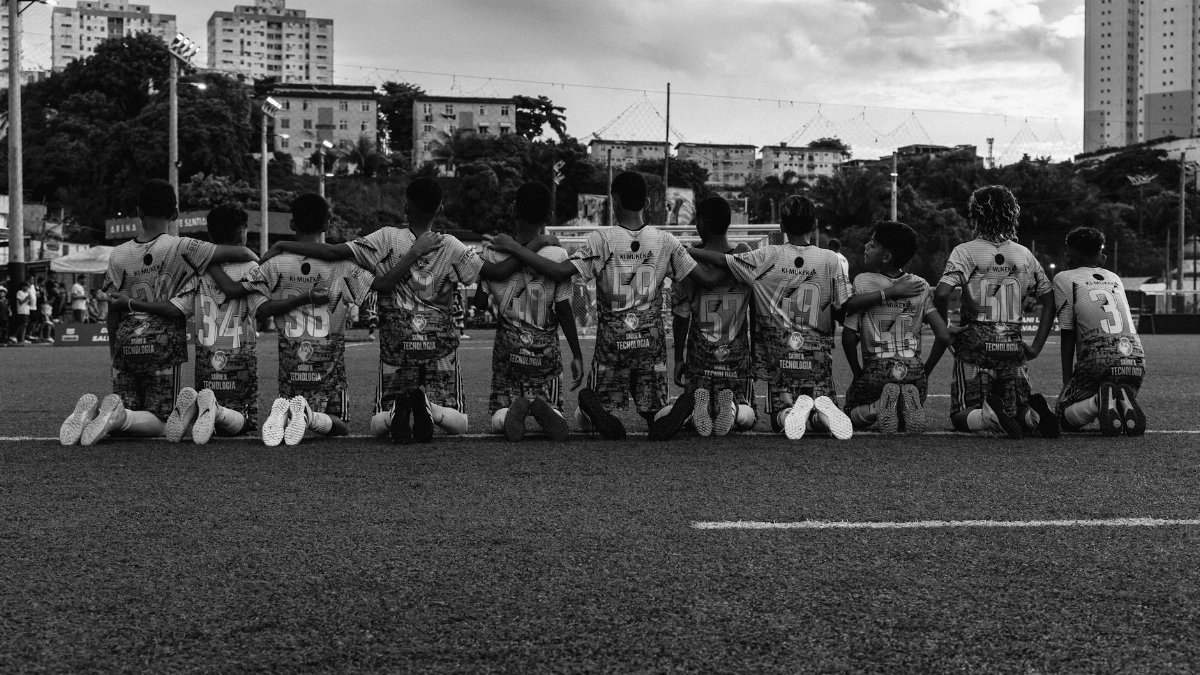 Black and white photo of a soccer team kneeling and embracing before a game on an urban field.
