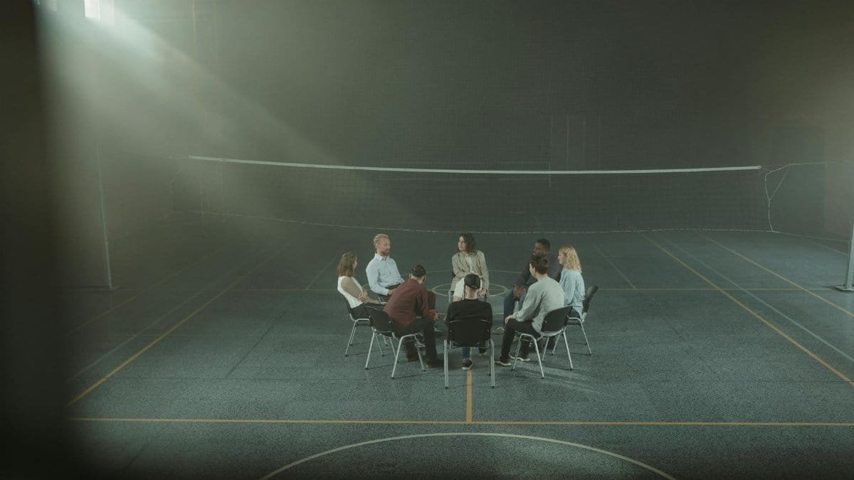 A group therapy session with participants seated in a circle inside a dimly lit gymnasium.