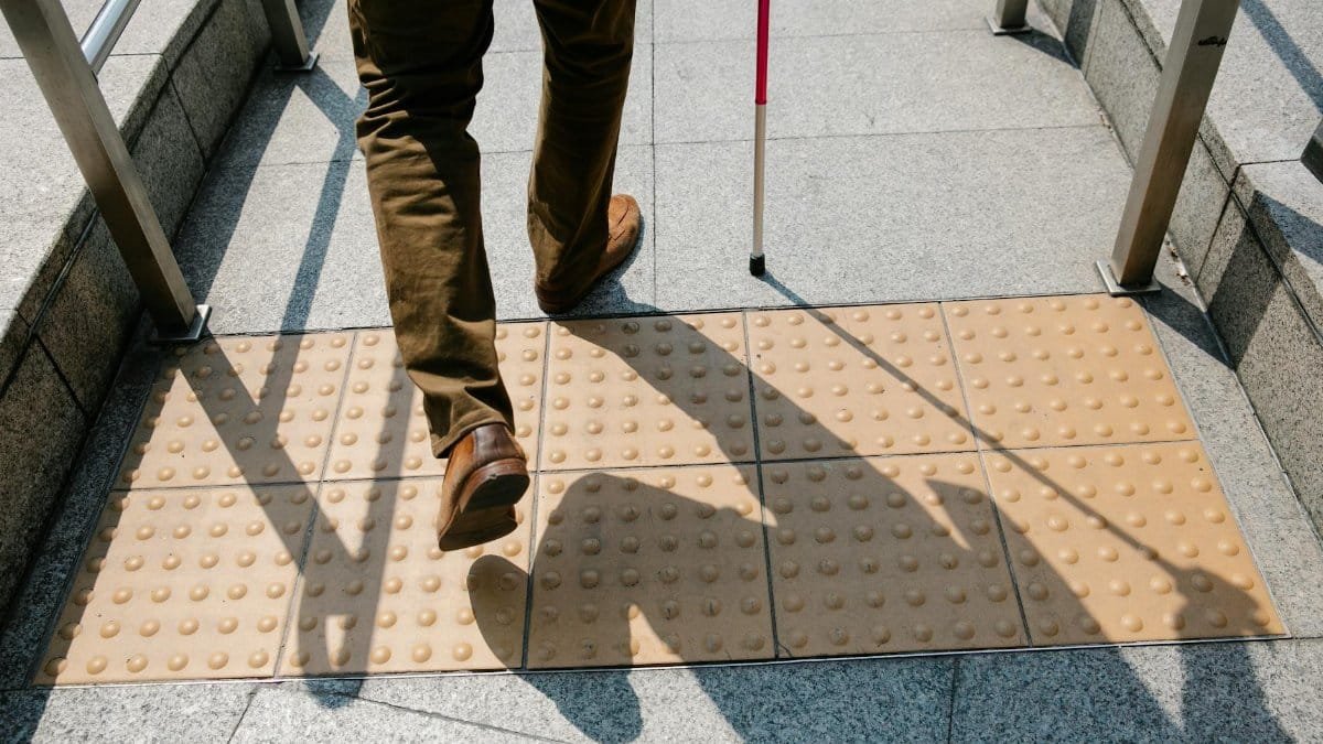 A person using a cane walks on tactile paving stones on a sunny day, emphasizing accessibility.