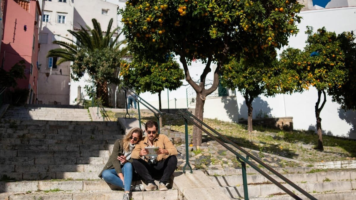 Couple sitting on steps with a map, surrounded by vibrant orange trees in a sunny urban setting.