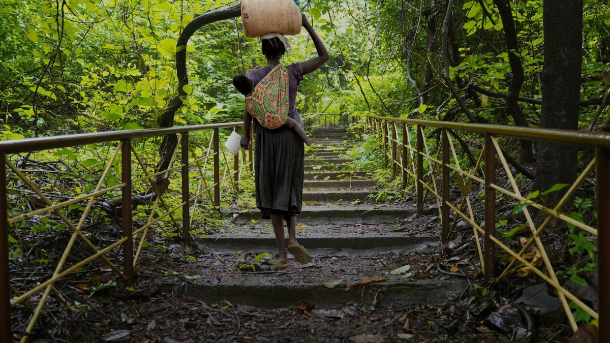 A woman carrying a child and a barrel ascends stone steps through lush forest greenery.