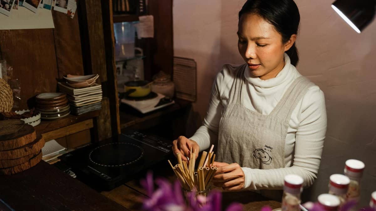 An Asian female bartender arranging straws in a cozy, rustic café setting during the evening.
