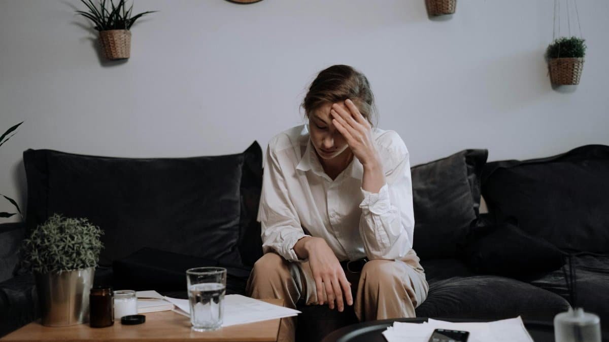 Woman experiencing stress and headache while sitting on a sofa at home, surrounded by papers.
