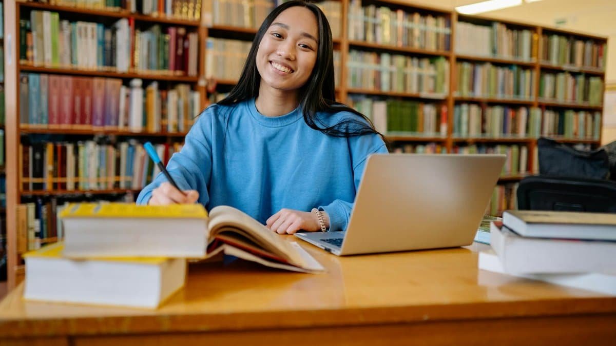 Smiling student studying in a library with books and laptop, showcasing education and focus.