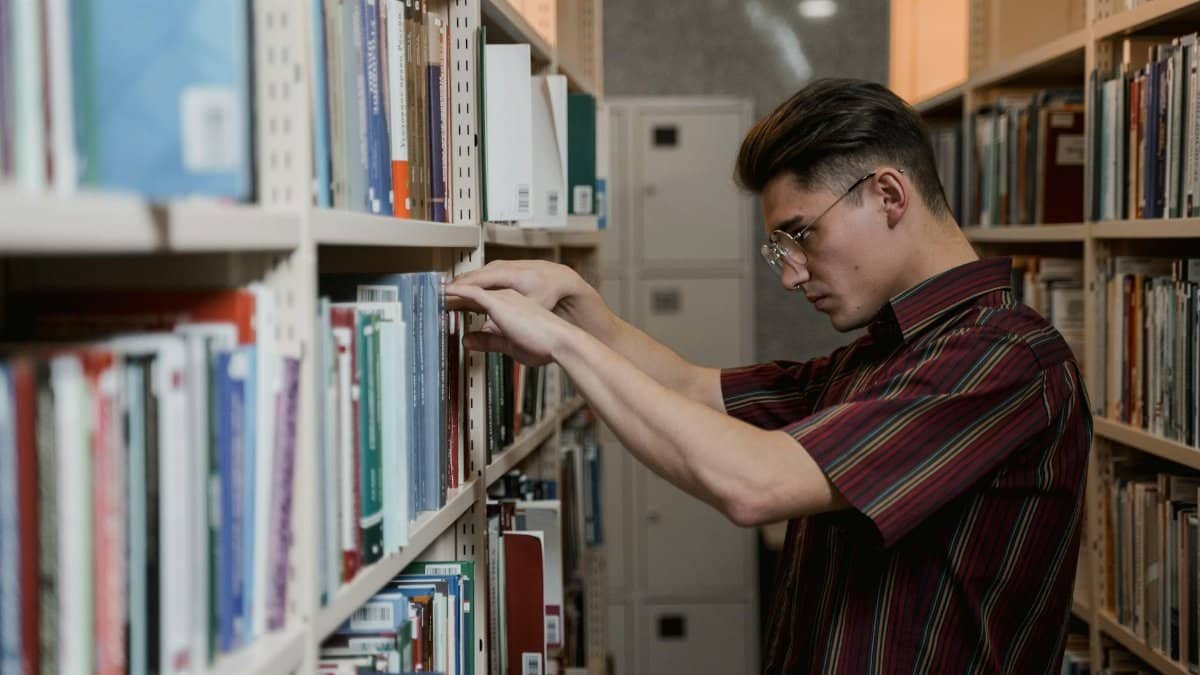 A young man in glasses searches library shelves for academic resources.