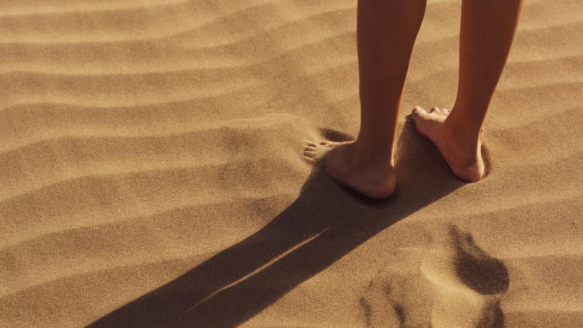 Close-up of a person standing barefoot on sunlit sand with visible footprints.