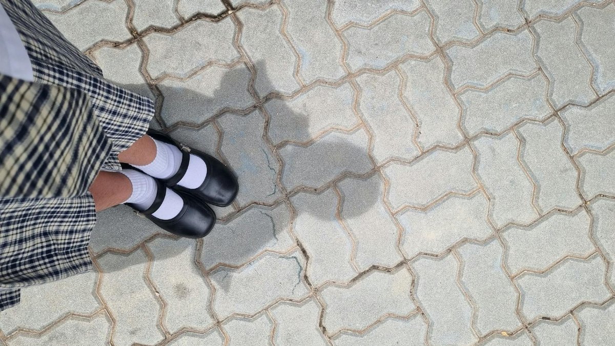 A woman in a plaid skirt and black shoes stands on a patterned pavement. Her shadow is visible.