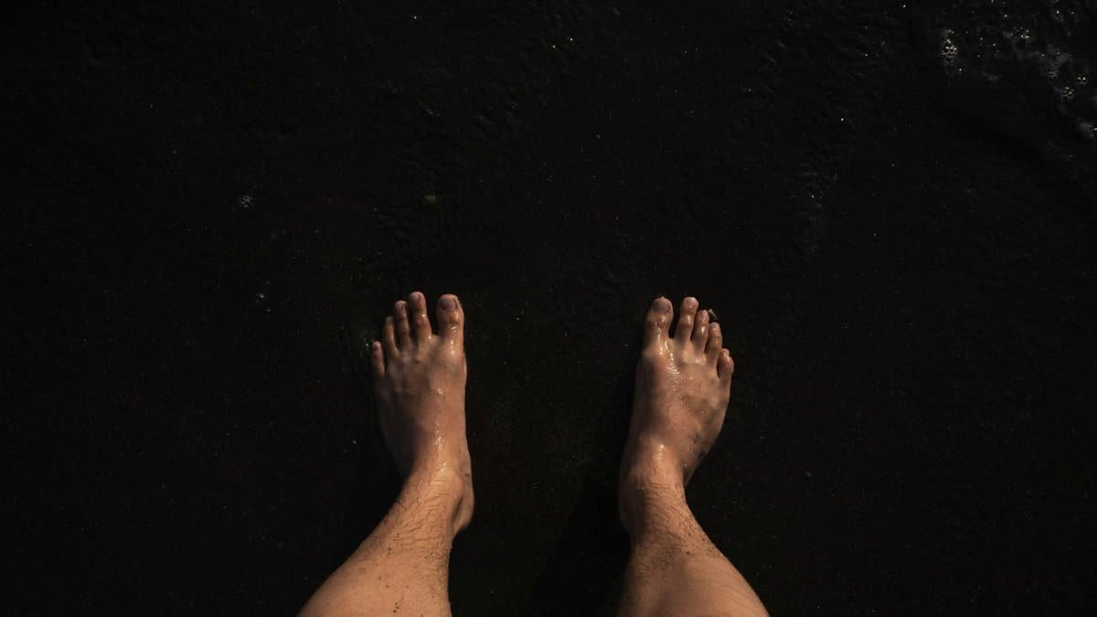View of a man's wet feet standing on dark sand at the beach.