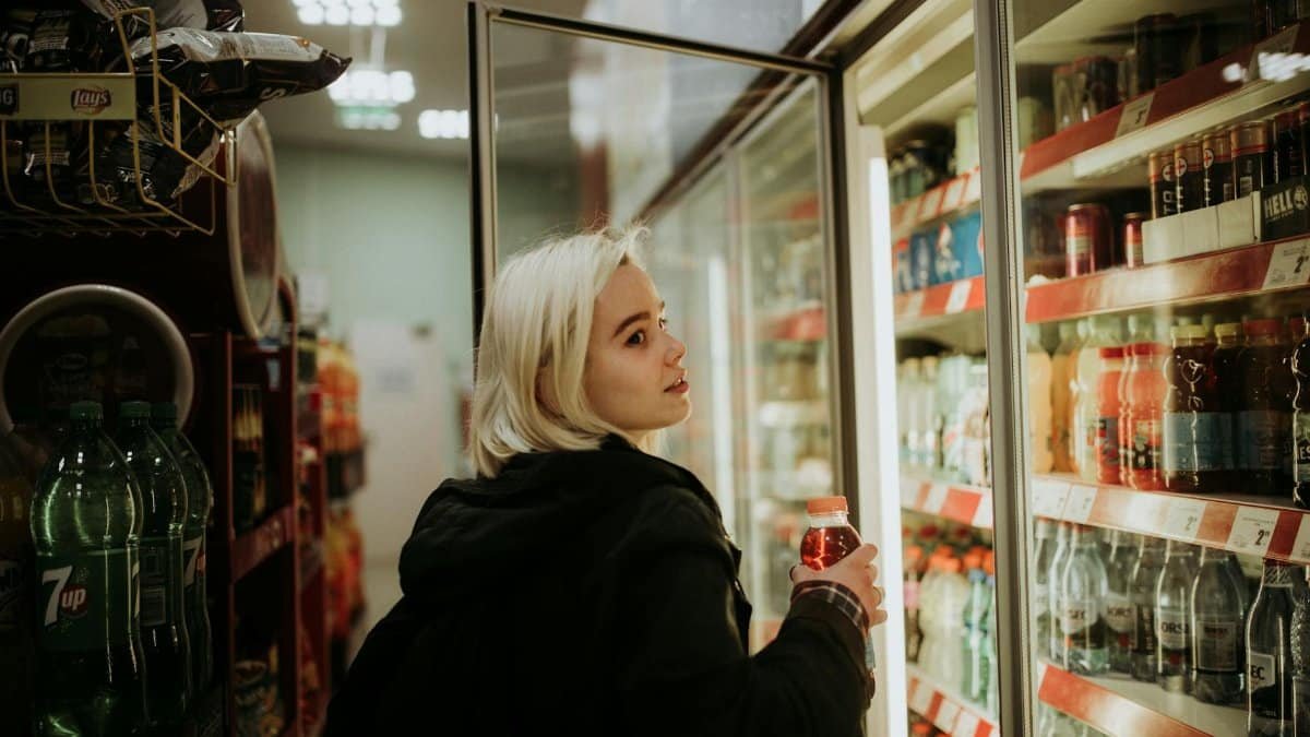 Blonde woman selecting a beverage from a refrigerated shelf at a convenience store.