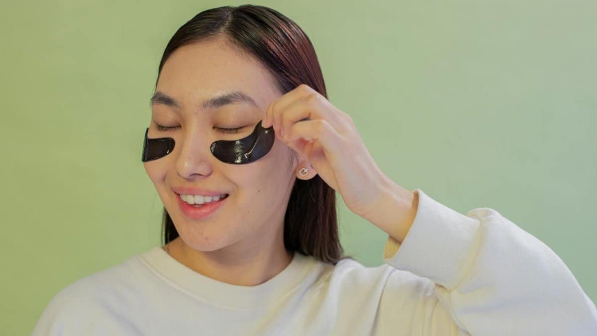 Asian teen girl smiling while applying black under-eye patches for skincare, green background.