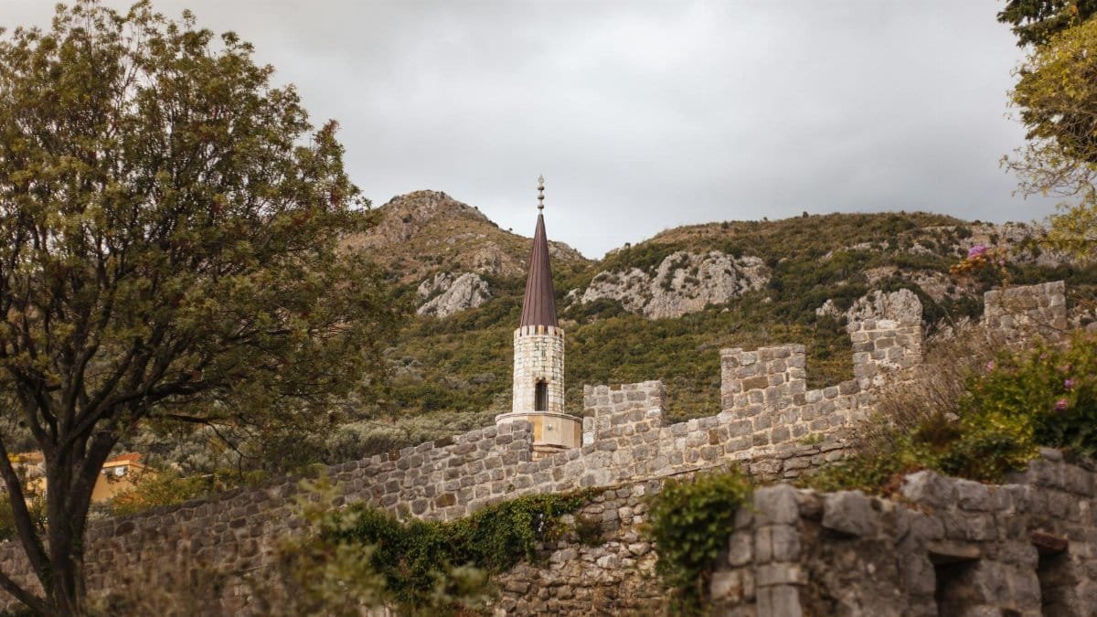 A medieval stone wall with a tower amidst lush greenery and mountains under a cloudy sky.