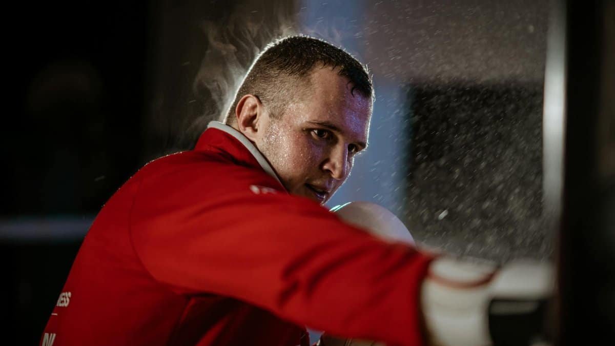 Strong male athlete in sportswear and boxing gloves practicing punches while looking forward in gymnasium