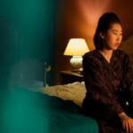 A woman in a robe sits thoughtfully on a bed in a warmly lit elegant bedroom.