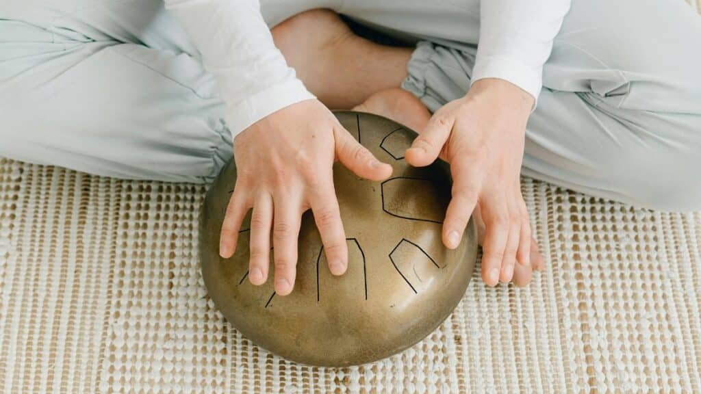 A woman sitting cross-legged playing a steel tongue drum indoors, enhancing peace and mindfulness.