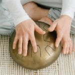 A woman sitting cross-legged playing a steel tongue drum indoors, enhancing peace and mindfulness.