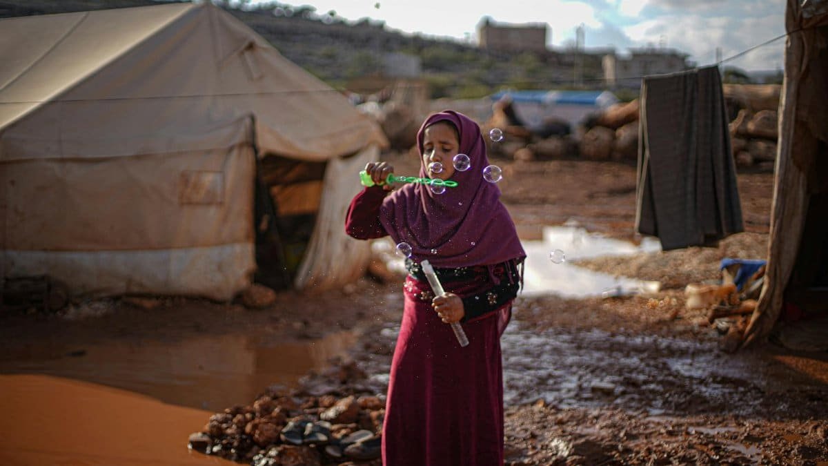 A girl in a Syrian refugee camp enjoys blowing bubbles amidst tents, capturing a moment of childhood joy.