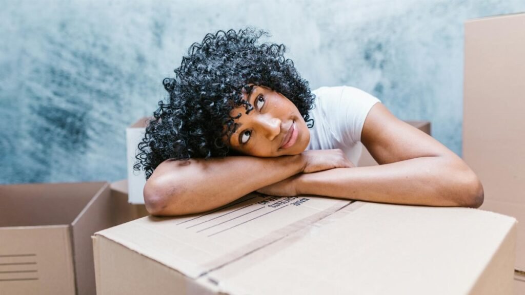 African American woman with curly hair resting on moving boxes indoors.