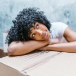 African American woman with curly hair resting on moving boxes indoors.