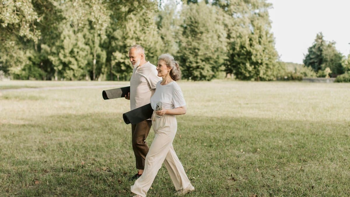 Elderly couple walking on grass carrying yoga mats, enjoying a sunny day outdoors.