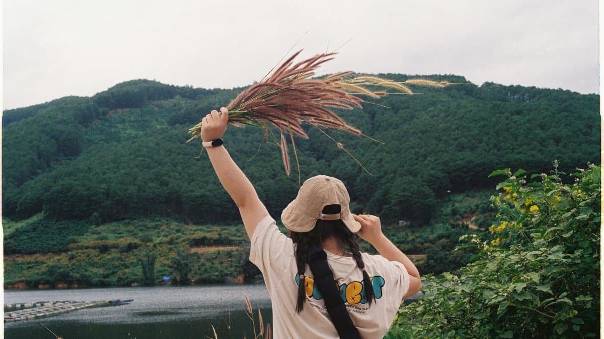 Girl with hat holds grass by a mountain lake, showcasing nature's beauty and tranquility.