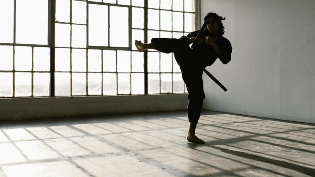 Martial artist in silhouette practicing a high kick in an industrial studio space.
