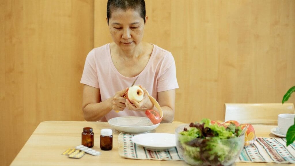 Middle aged ethnic female with short hair in t shirt peeling fresh apple and preparing salad while sitting at table with veggies and various medicine during remission