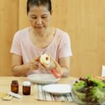 Middle aged ethnic female with short hair in t shirt peeling fresh apple and preparing salad while sitting at table with veggies and various medicine during remission
