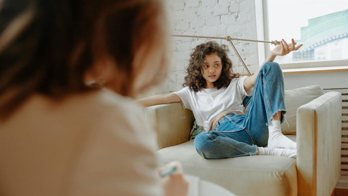 A teenager with curly hair receives counseling in a modern indoor setting, discussing mental health.