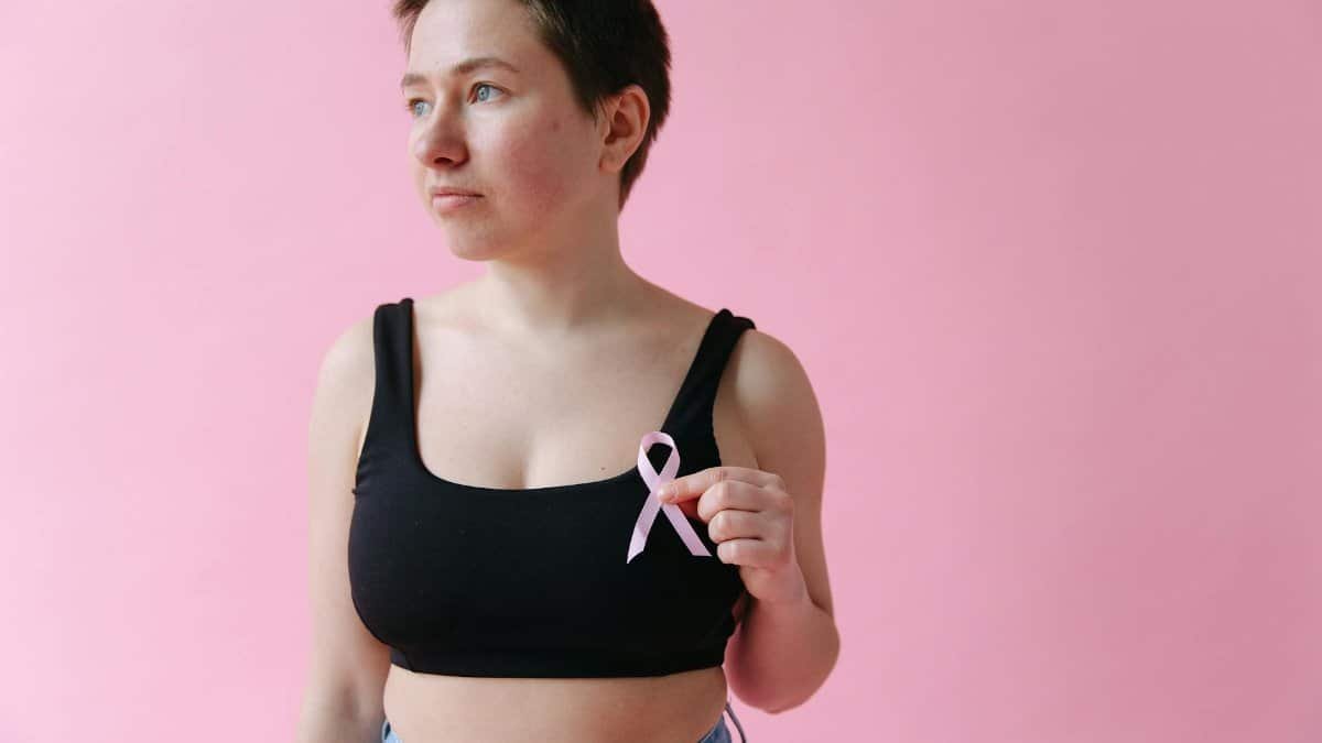 Woman holding a pink ribbon symbolizing breast cancer awareness, posing against a pink background.