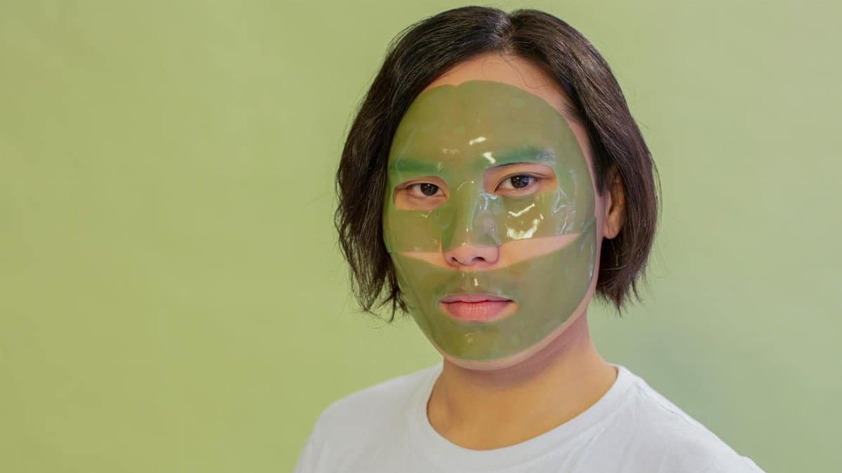 Adult wearing a green facial mask on a light green background, promoting skincare and wellness.