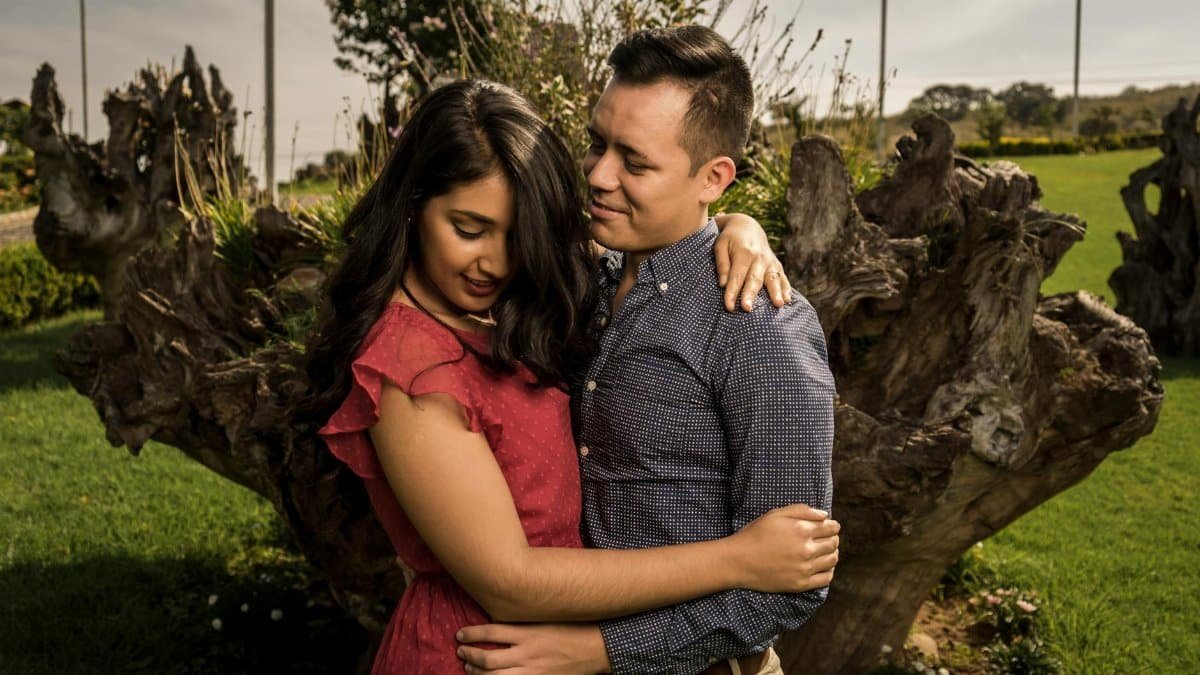 A loving couple embraces in a sunny outdoor setting surrounded by nature.