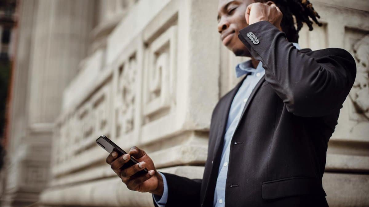 Low angle of happy young black guy in formal wear browsing mobile phone and enjoying music in true wireless earphones while standing on street near modern building