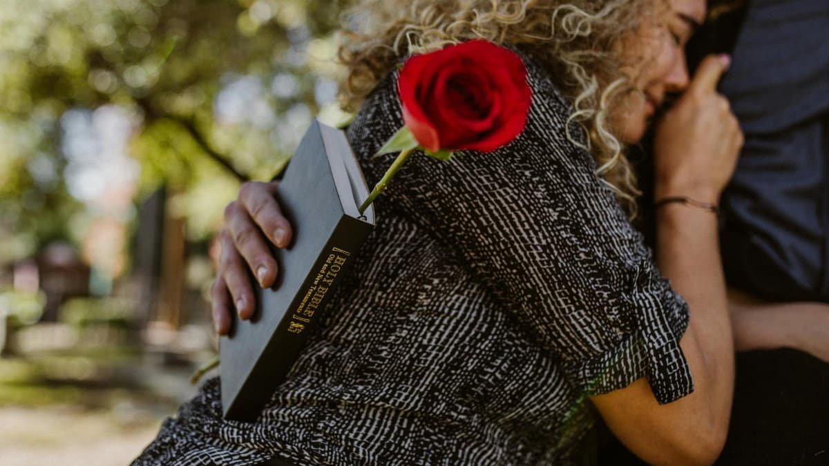 A woman embraces a person outdoors, holding a holy Bible and a red rose, reflecting deep emotion.