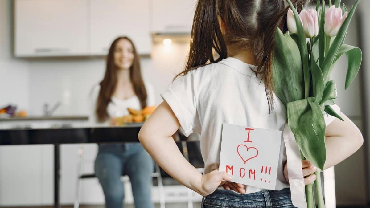 Back view of little girl with ponytails in casual clothes standing against mother with hands behind back and holding bouquet of tulips and drawing I love mom on kitchen