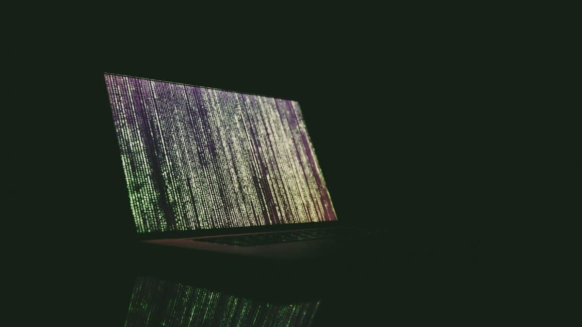 A laptop displaying green and purple code in a dark room, reflecting on a surface.