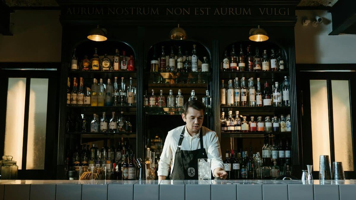 A young male bartender mixes a cocktail behind a well-stocked bar counter.