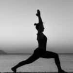 Grayscale silhouette of a woman practicing yoga on a beach at sunset.