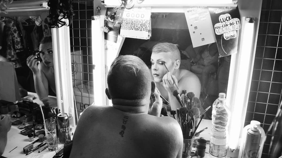 Drag queen applying makeup in dressing room, showcasing artistry and transformation.