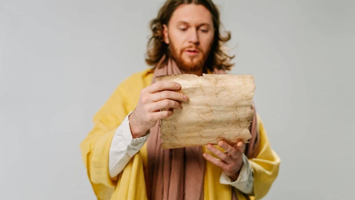Man with long hair reads an ancient scroll. Historical attire suggests a religious or scholarly theme.