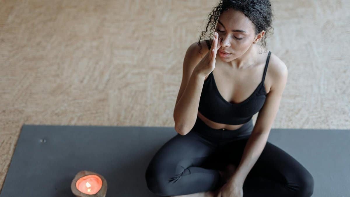 A woman practicing meditation on a yoga mat, focusing with eyes closed next to a candle indoors.